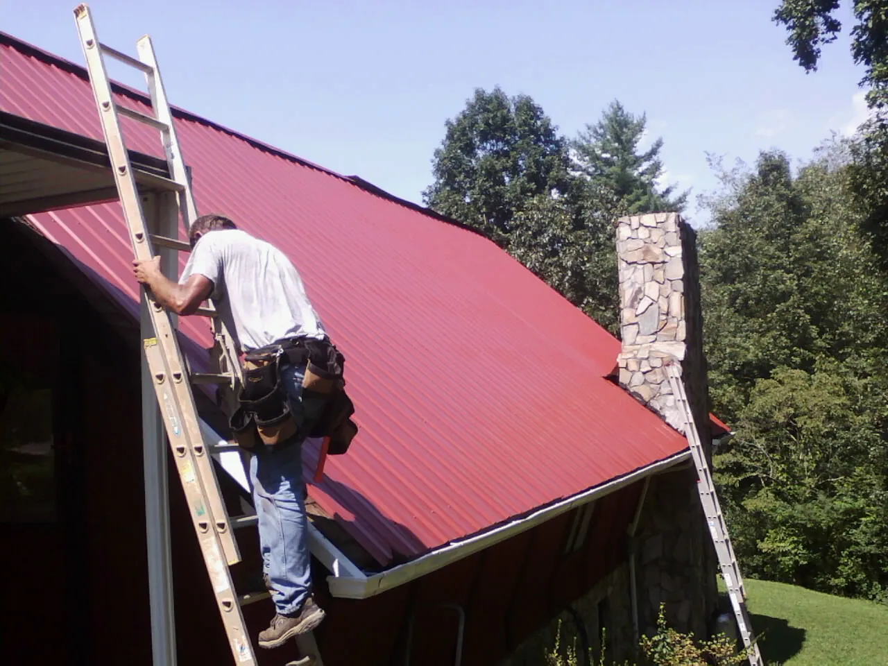 Roofer on a ladder beside a red metal roof and stone chimney — Soffit & Fascia Repair by Roofing Brothers in Sumterville